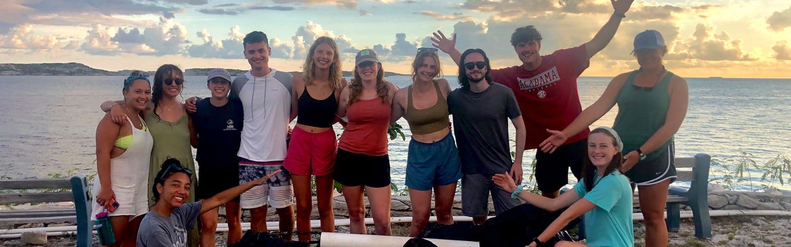 A group of students pose for the camera while standing on a beach overlooking the ocean that is reflecting the sunset. 